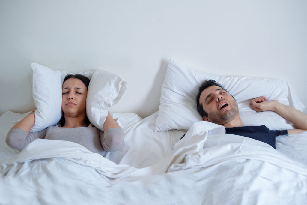Sleep Disturbances, woman covering ears with a pillow due to man snoring, man is sleeping with mouth open